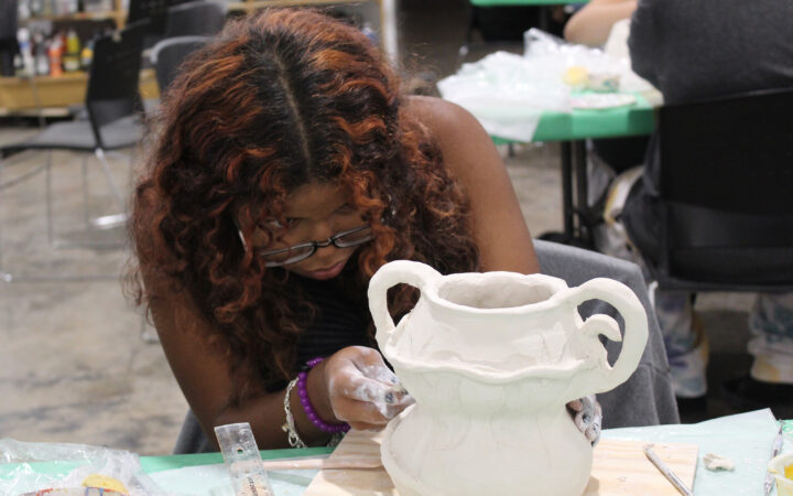 A photograph of a young teenager working diligently on a cermaic pot. She looks to be shaping the curve in the pot.