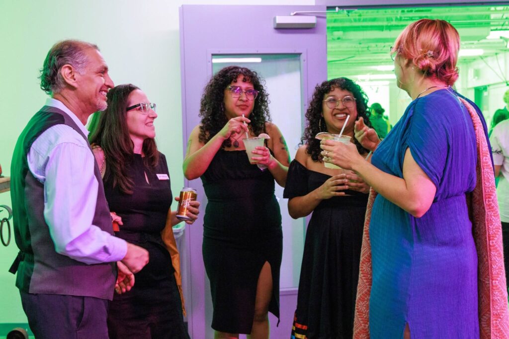 An image of five individuals smiling at a party while talking and holding beverages.