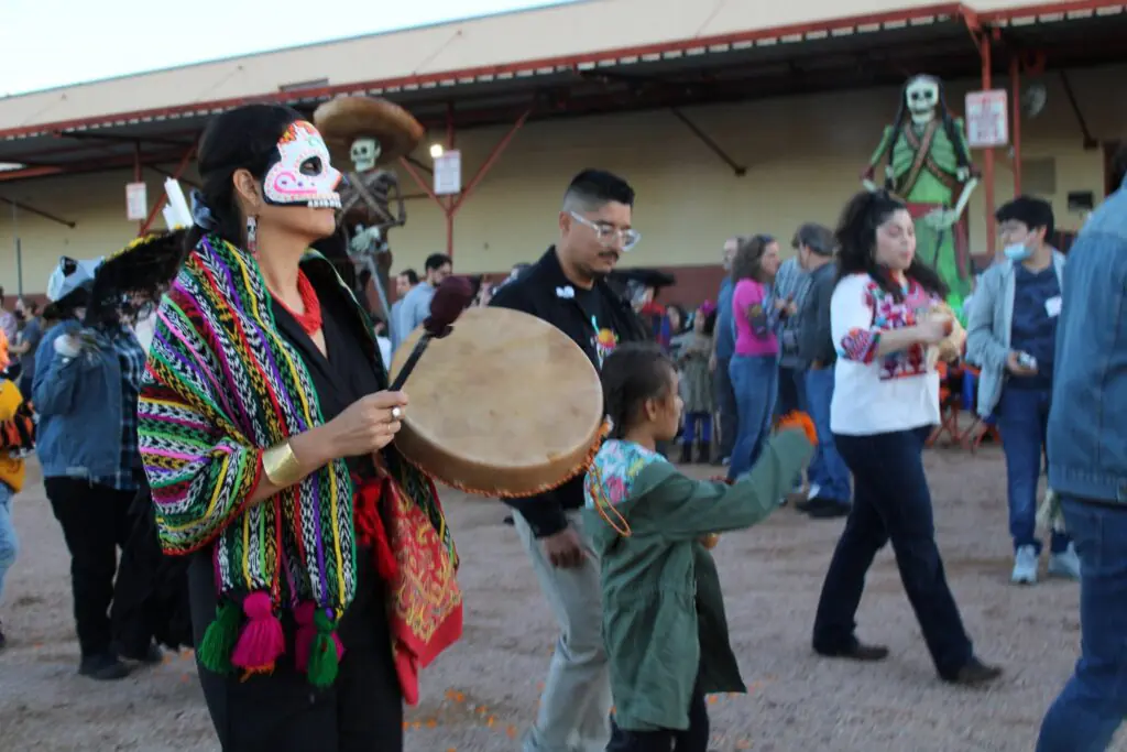 This is an image of people walking in a street procession. Some are painted as skulls, and some hold various instruments like drums.